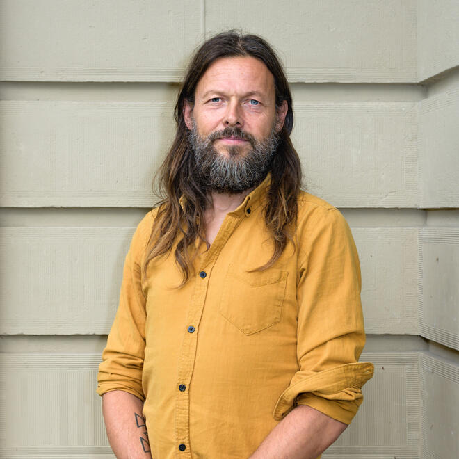 Foto van Joost van Pagée, een man met lang haar en een baard. Hij zit op een steen en draagt een blauw shirt, blauwe schoenen en een blauwe spijkerbroek