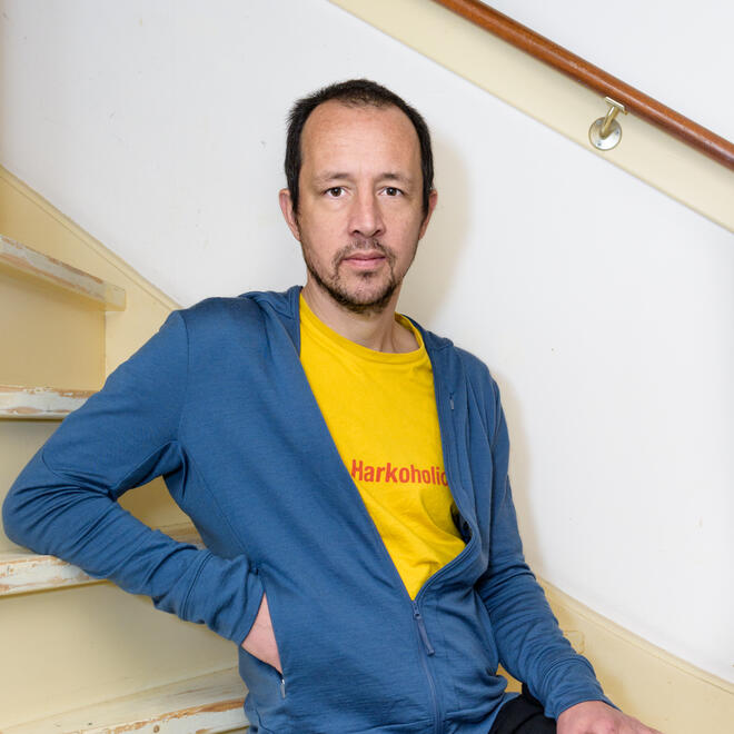Foto van Floris van Bergeijk die op een bankje zit. Hij heeft donker kort haar en draagt een blauw vest met een rood shirt er onder. Hij zit op een bankje buiten